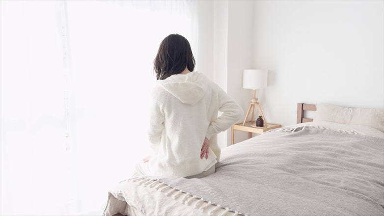 朝起きてベッドに腰掛け、腰の違和感を感じている女性