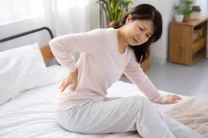 朝の起き上がりで腰が痛い50代女性。ベッドから起き上がろうとし、腰に手を当てている様子。