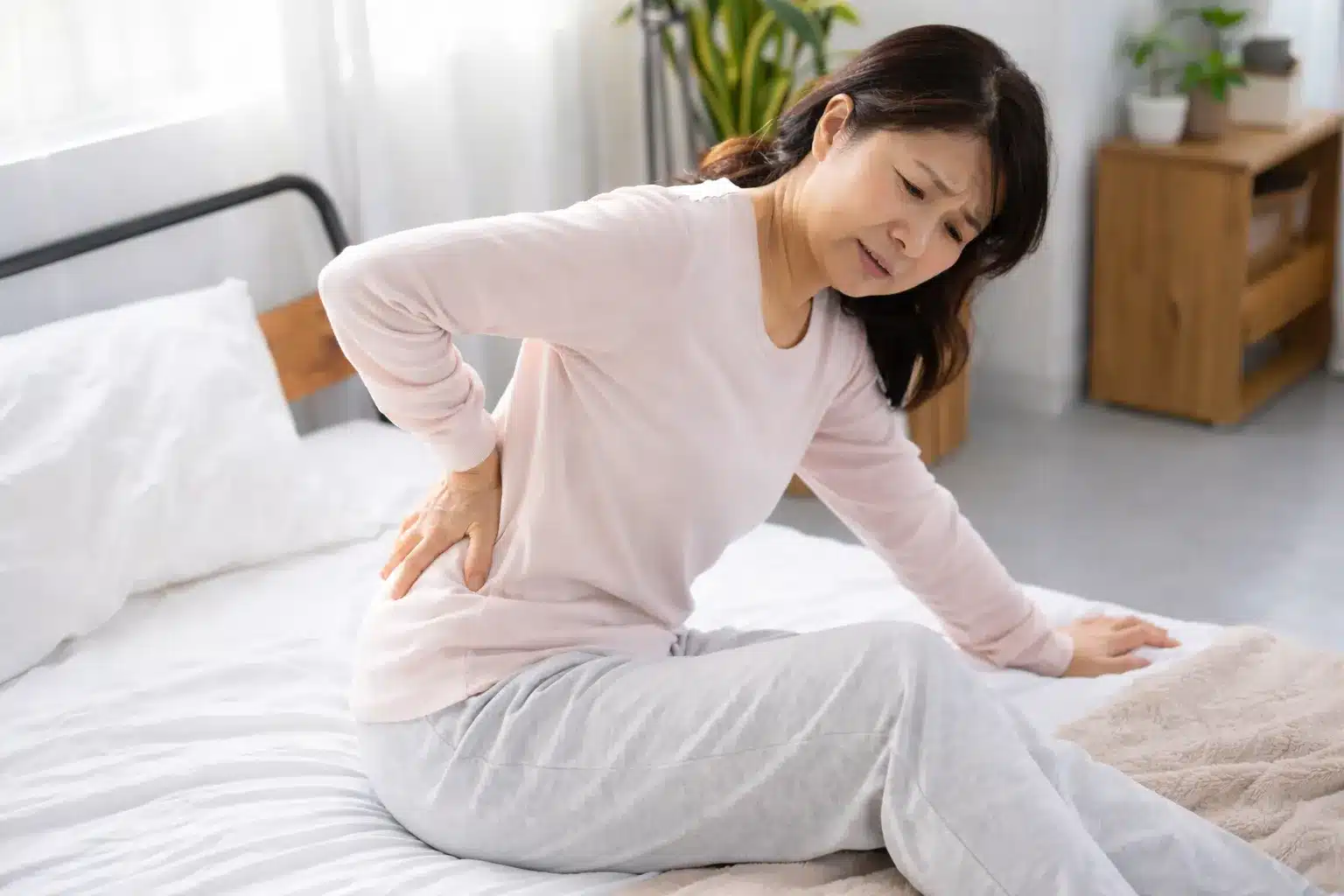 朝の起き上がりで腰が痛い50代女性。ベッドから起き上がろうとし、腰に手を当てている様子。